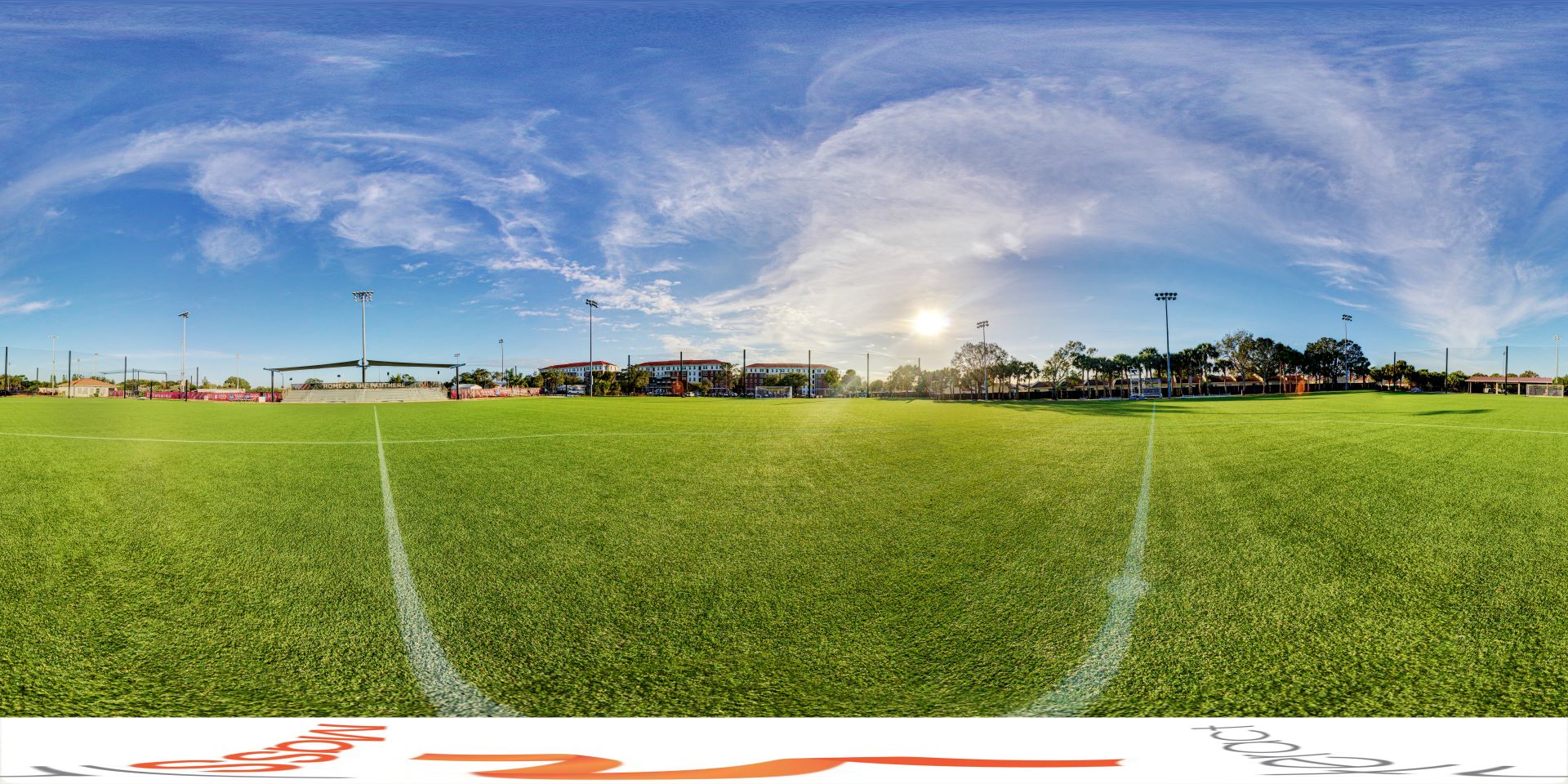 A panoramic view of Rick Stottler Field at Florida Tech, showcasing the green soccer field, bleachers, and buildings in the background under a clear blue sky with some clouds